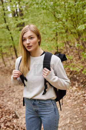 Foto de Retrato de joven viajera rubia con mochila senderismo en el bosque de otoño y de pie en el camino - Imagen libre de derechos