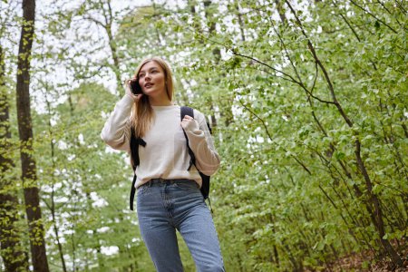 Photo of blond pretty woman traveler with backpack talking by phone walking in forest path