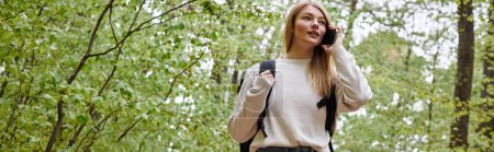 Photo of blond pretty woman traveler with backpack talking by phone walking in forest path, banner
