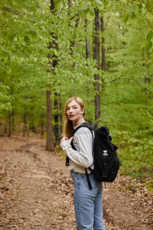 Photo for Side view shot of blond pretty woman traveler with backpack looking for direction walking in forest - Royalty Free Image