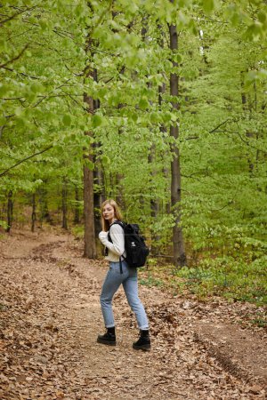 Young blonde female hiker wearing backpack crossing the forest path walking in woods
