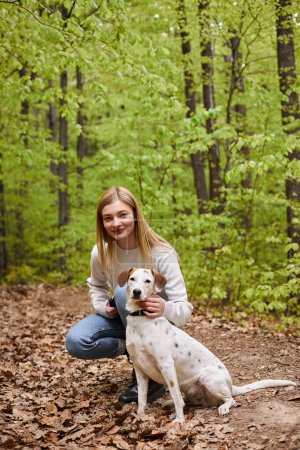 Foto de Sonriente chica excursionista interactuando con su mascota mirando a la cámara mientras camina resto con vista al bosque - Imagen libre de derechos