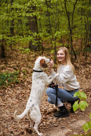 Téléchargez les photos : Souriant randonneuse fille interagissant avec son entraînement pour animaux de compagnie pendant la randonnée repos avec vue sur la forêt - en image libre de droit