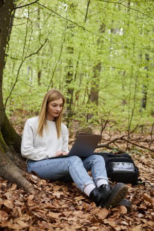 Junge Wanderin mit Laptop an den Beinen, Pullover, Jeans und Wanderschuhen im Wald sitzend
