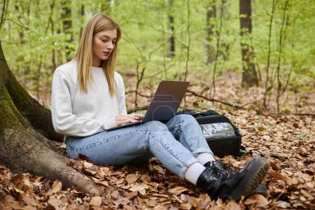 Blonde Wanderin mit Laptop an den Beinen arbeitet ferngesteuert, während sie im Wald unterwegs ist