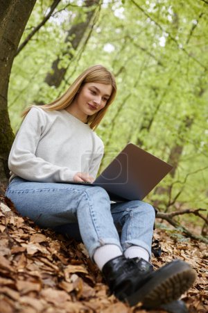 Lächelnde blonde Wanderin mit Laptop an den Beinen, die ferngesteuert arbeitet, während sie unterwegs im Wald sitzt