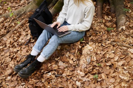 Cropped view of woman hiker with laptop on her knees working remote while sitting in forest on trip