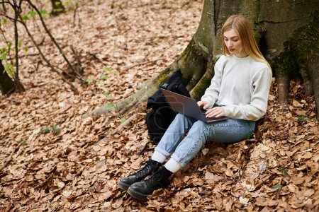 Young calm woman hiker with laptop on her knees working remote while sitting in forest on trip