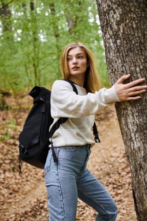 Foto de Relajada mujer rubia senderismo y caminar en el bosque verde tocando árboles girando en dirección - Imagen libre de derechos