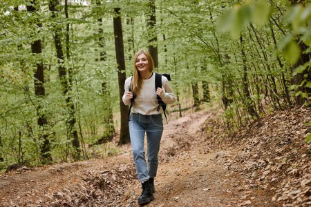 Foto de Feliz senderismo mujer rubia con suéter y mochila caminando en el paisaje del bosque en los bosques - Imagen libre de derechos