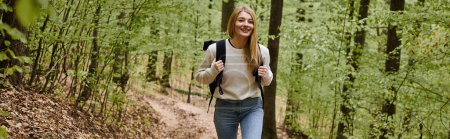 Happy hiking blonde woman wearing sweater and backpack walking in forest scenery in woods, banner