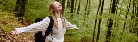 Photo for Smiling relaxed blonde woman hiker wearing sweater and backpack with arms  open in forest, banner - Royalty Free Image