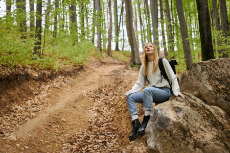 Curious blonde female hiker wearing backpack, resting on rocks, sitting in deep woods