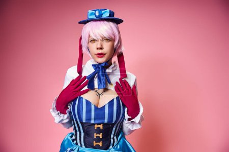 beautiful stylish female cosplayer in blue hat and vivid dress looking at camera on pink backdrop