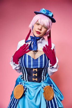 beautiful stylish female cosplayer in blue hat and vivid dress looking at camera on pink backdrop
