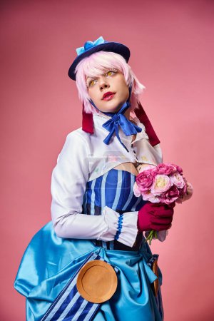 Photo for Fashionable female cosplayer in vivid attire holding pink flowers and looking away on pink backdrop - Royalty Free Image