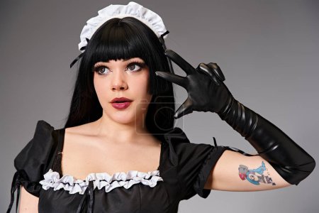 alluring young cosplayer in black maid costume gesturing on gay background and looking away