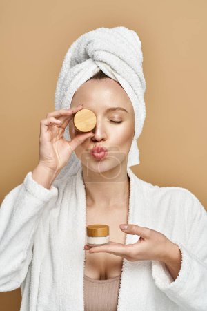 Une femme séduisante avec une serviette sur la tête tenant gracieusement un pot de crème, soulignant sa routine de beauté naturelle.