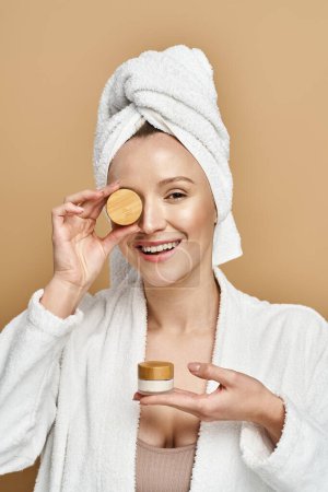 An attractive woman with a towel on her head holds a jar of cream, showcasing her natural beauty routine.