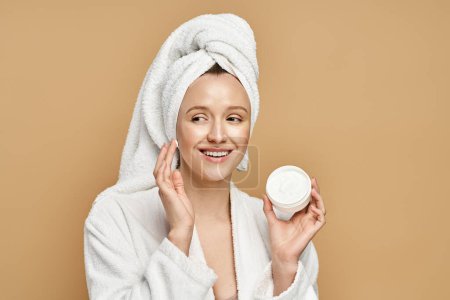 A woman with a towel on her head holds a jar of cream, showcasing her beauty routine in a serene setting.