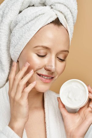 Une femme magnifique avec une serviette enroulée autour de sa tête est vue tenant un pot de crème, prêt à l'appliquer sur sa peau.