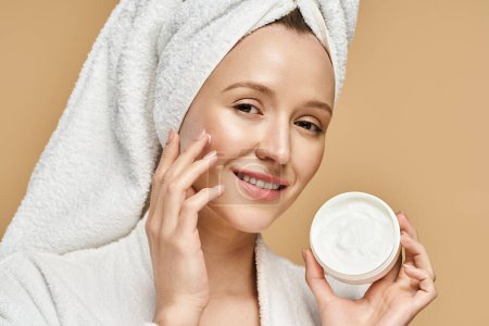 An attractive woman with a towel on her head gracefully holds a jar of cream, exuding natural beauty and elegance.