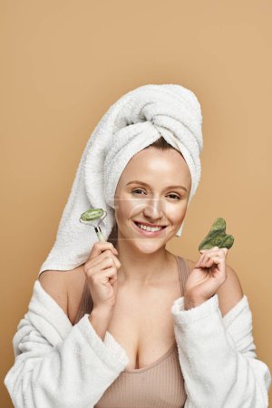A woman exuding natural beauty wears a towel turban while delicately holding a face roller in a serene pose.