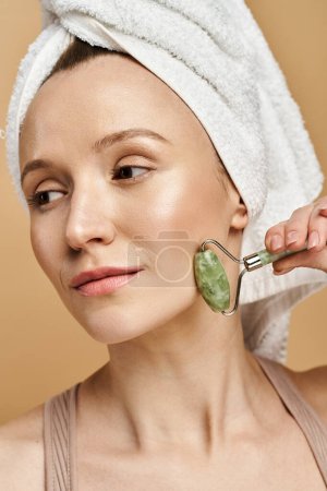 Photo for An attractive woman with a towel on her head, artfully holding face roller. - Royalty Free Image