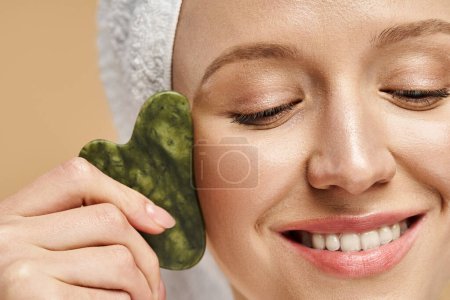 A serene and attractive woman with a towel wrapped around her head holds a vibrant gua sha in her hand.