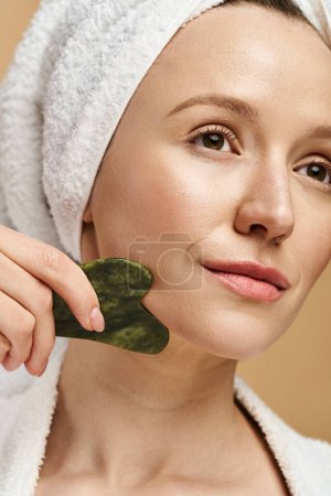 A woman, showcasing natural beauty, holds a gua sha while a towel rests on her head.