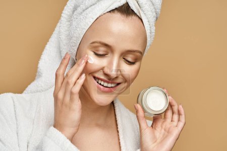 Una mujer atractiva con una toalla en la cabeza aplicándose crema en la cara.
