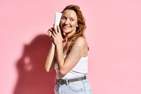 A graceful woman in a white tank top holding cream with focus and ease, embodying natural beauty and modern connectivity.