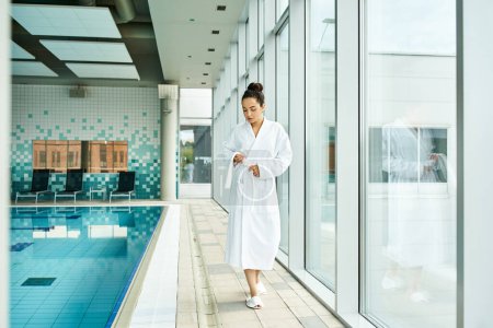 Photo for A young, beautiful brunette woman in a bathrobe standing beside an indoor swimming pool. - Royalty Free Image