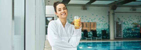 A brunette woman in a luxurious bathrobe leisurely sips orange juice by an indoor spa with a swimming pool.