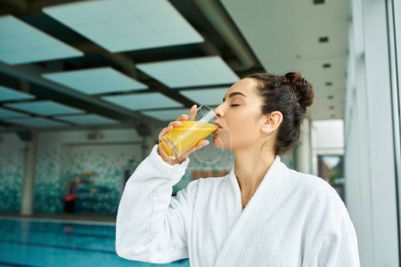 A young, beautiful brunette woman luxuriously savoring a refreshing glass of orange juice in an indoor spa by a swimming pool.
