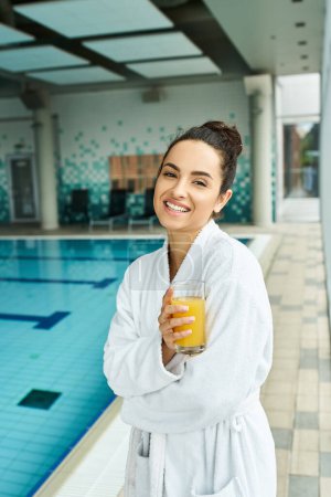 A young brunette woman relaxes in an indoor spa, savoring a glass of orange juice in her bathrobe by the swimming pool.