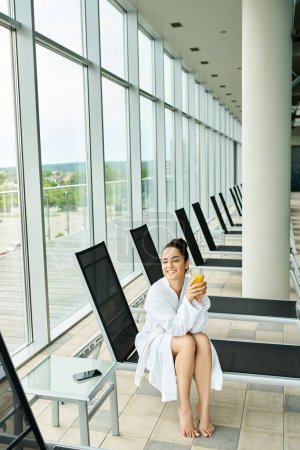 A young, beautiful brunette woman sits serenely on a bench in an indoor spa with a swimming pool, clad in a luxurious bathrobe.