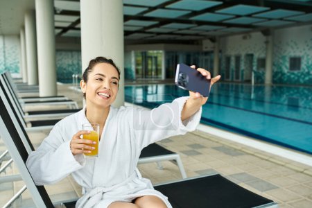 Photo for A young brunette woman in a bathrobe enjoying a drink and scrolling on her cell phone in an indoor spa by a swimming pool. - Royalty Free Image
