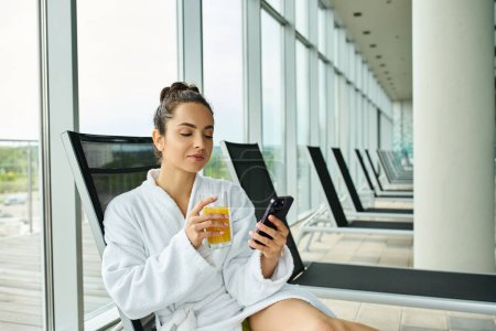 Une jeune femme brune assise sur une chaise près d'un spa intérieur, tenant un téléphone portable.