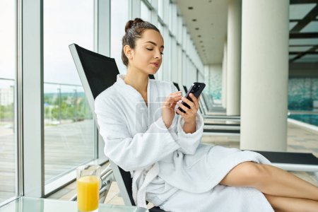 A young, brunette woman is seated in a luxurious bathrobe, engrossed in her cell phone in an indoor spa with a swimming pool.