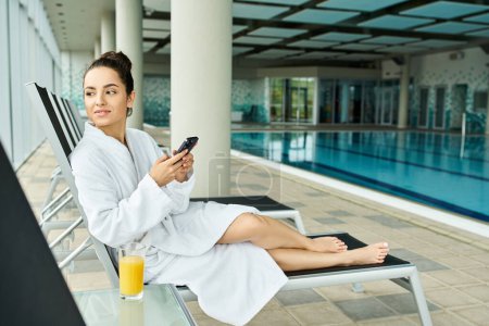 A young, beautiful brunette woman in a bathrobe is sitting on a lounge chair by an indoor swimming pool.