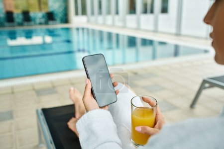Foto de Una joven y hermosa mujer sostiene un teléfono celular junto a una piscina cubierta, capturando recuerdos. - Imagen libre de derechos