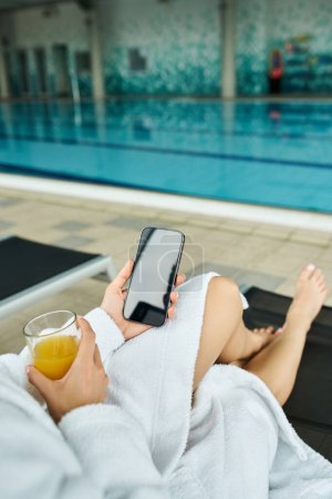 Téléchargez les photos : Une jeune femme brune allongée à côté d'une piscine intérieure, en utilisant paisiblement son téléphone portable. - en image libre de droit