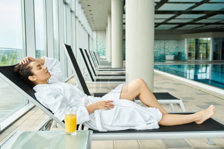 Una joven y hermosa morena está descansando en un sillón junto a una piscina cubierta, disfrutando de la relajación.
