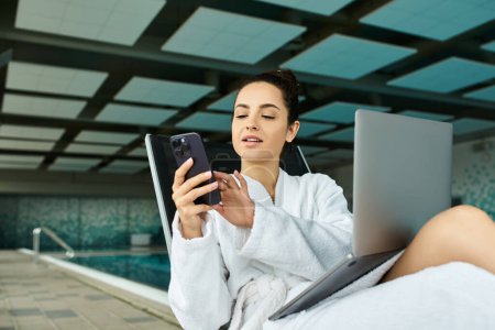 Une jeune et belle brune se détend dans un peignoir au bord d'une piscine thermale intérieure, concentrée sur son téléphone portable.