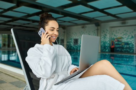 Eine junge, schöne brünette Frau im Bademantel, die in einem Indoor-Wellnessbereich mit Swimmingpool mit einem Handy telefoniert.