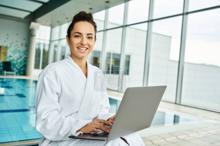 Eine junge, schöne brünette Frau im Bademantel entspannt am Pool, in ihren Laptop vertieft.
