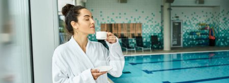 A young brunette woman enjoys a peaceful morning, sipping coffee in a luxurious bathrobe by an indoor spa pool.