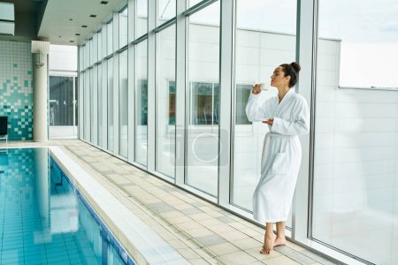 Téléchargez les photos : Une jeune, belle femme brune en peignoir blanc se tient gracieusement à côté d'une piscine intérieure. - en image libre de droit
