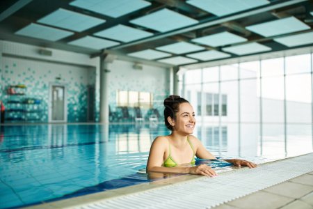 Téléchargez les photos : Une jeune, belle femme brune se détend dans un bikini vert dans une piscine spa intérieure, respirant élégance et tranquillité. - en image libre de droit